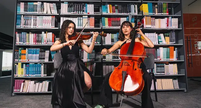Dos jovenes mujeres en la derecha una tocando un violonchelo y la de la izquierda tocando un violin 