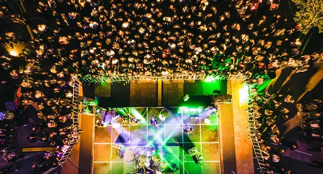 Vista aérea nocturna de un concierto al aire libre en la Anáhuac Querétaro. Una multitud rodea un escenario iluminado con luces verdes y moradas.