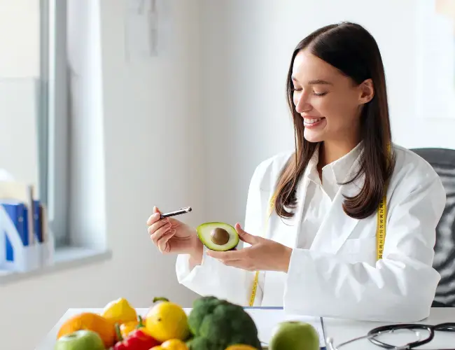 Una mujer con bata realizando actividades que hace un nutriólogo, escribiendo en un formulario con frutas y verduras en su escritorio