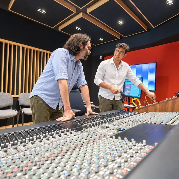 Dos adultos hombres platicando dentro del estudio de grabación uno esta tocando el sistema de audio el otro escucha. 